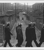 The greetings card oshowing a young woman returning home at lunchtime from her job at Hintons, the grocers. Police line the alleyway down which she is walking and a line of marching police officers pass the end of the alleyway.